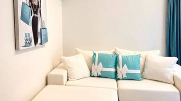 A white sofa with teal and white cushions, a framed picture on the wall, and a teal curtain.