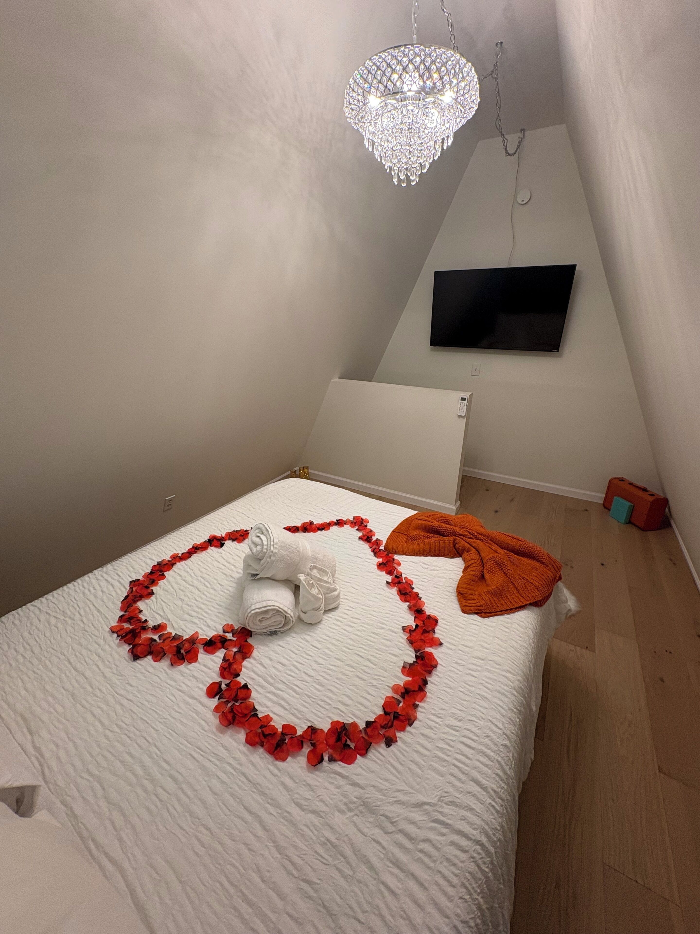 A bedroom with a bed, a chandelier, a flat-screen TV, and a heart-shaped decoration made of red flowers and white towels.