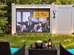 Una pantalla de cine al aire libre con 'THE WOLF OF WALL STREET' en exhibición, un cuenco de palomitas de maíz, una copa de whiskey y una vela sobre una mesa.