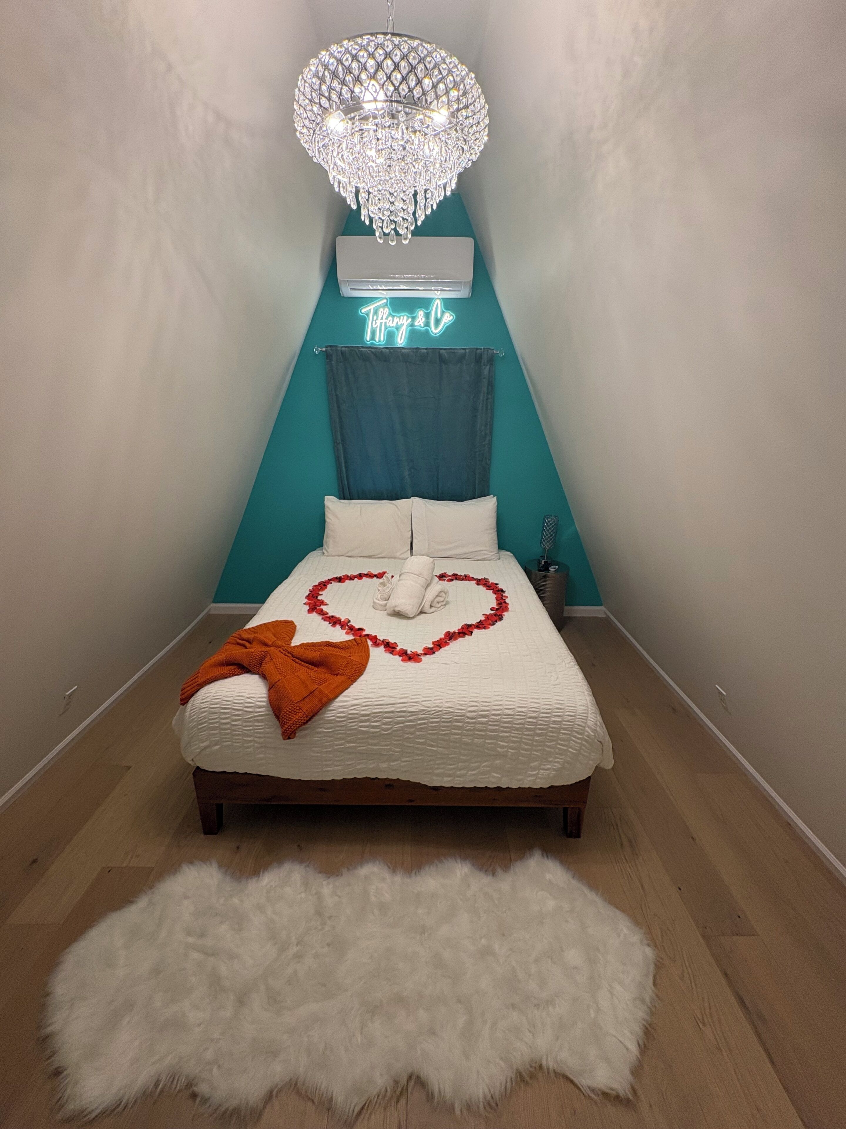 A bedroom with a bed, a chandelier, a neon sign, and a fluffy rug.