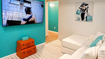 Un salon moderne avec un mur d’accent de couleur turquoise, un téléviseur à écran plat, un canapé blanc et un tableau représentant une femme tenant des sacs de courses.