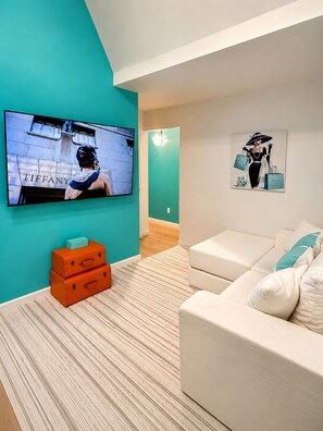 Ein modernes Wohnzimmer mit einer Wand in Türkis, einem Flachbildfernseher, einem weißen Sofa und einem Gemälde einer Frau mit Einkaufstüten.