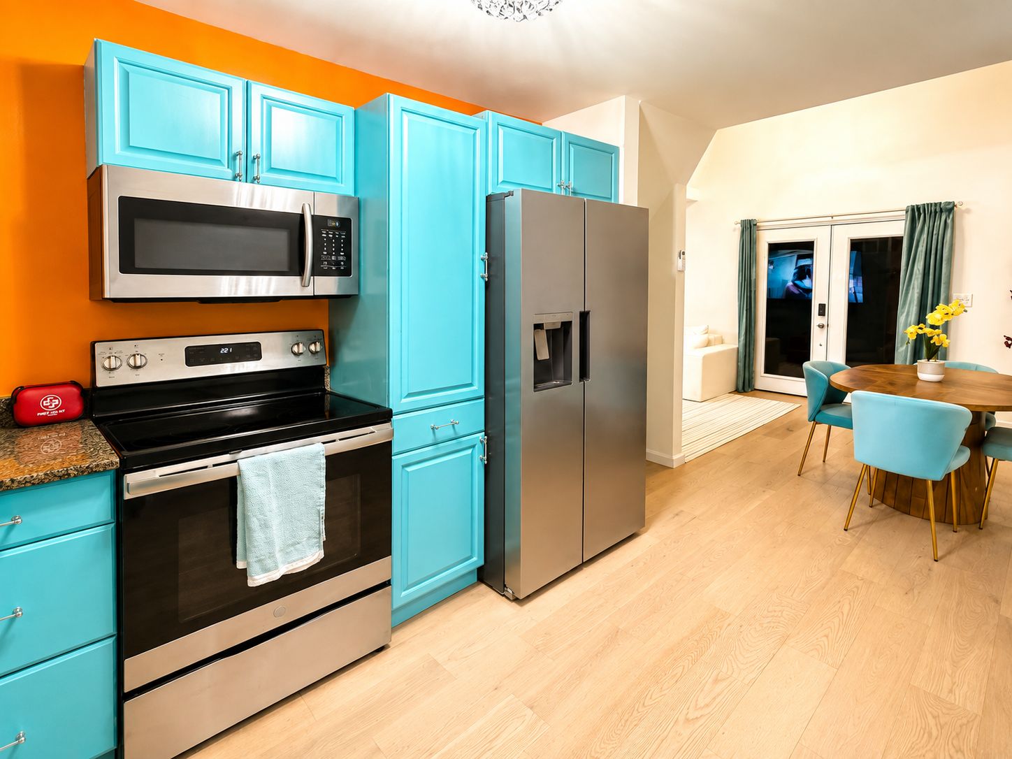 Une cuisine avec des armoires de couleur turquoise, un four à micro-ondes en acier inoxydable, un réfrigérateur et une table à manger en bois avec des chaises.