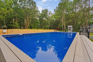 A rectangular swimming pool with a wooden deck surrounding it, set in a wooded area with trees and a small building in the background. - Pool, Hot Tub, Sauna, Game Room, Luxury and More! (Albrightsville)