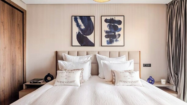 Una camera d'albergo con un letto grande, due cuscini con scritte, un comodino, una sveglia a muro e arte astratta sulla parete.