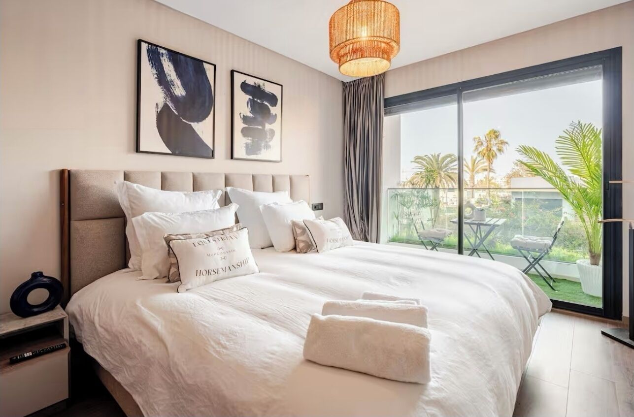 Un dormitorio de hotel con una cama grande, una mesita de noche, una ventana con vistas a palmeras y obras de arte en la pared.