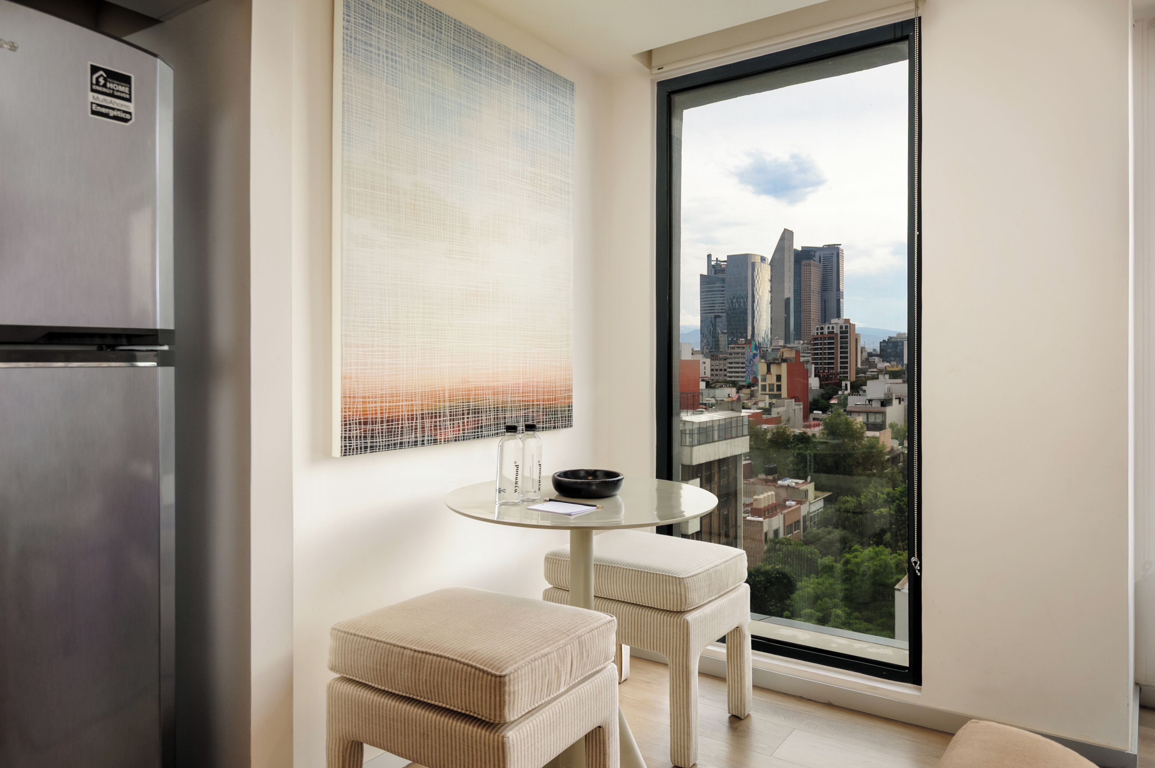 A modern kitchen with a stainless steel refrigerator, a round dining table with two cushioned chairs, and a large window offering a cityscape view.
