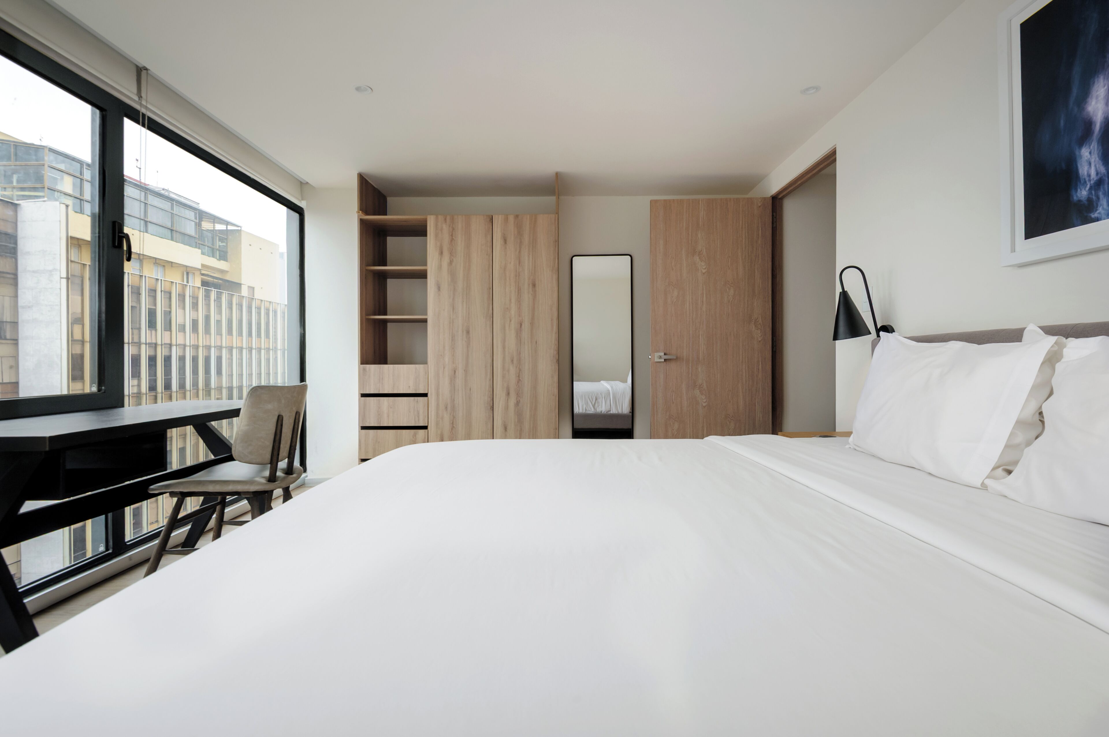A modern hotel room with a large bed, a desk, and a chair. There is a view of buildings outside the window.