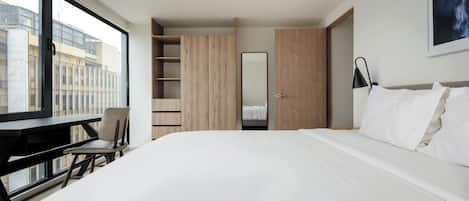 A modern hotel room with a large bed, a desk, and a chair. There is a view of buildings outside the window.