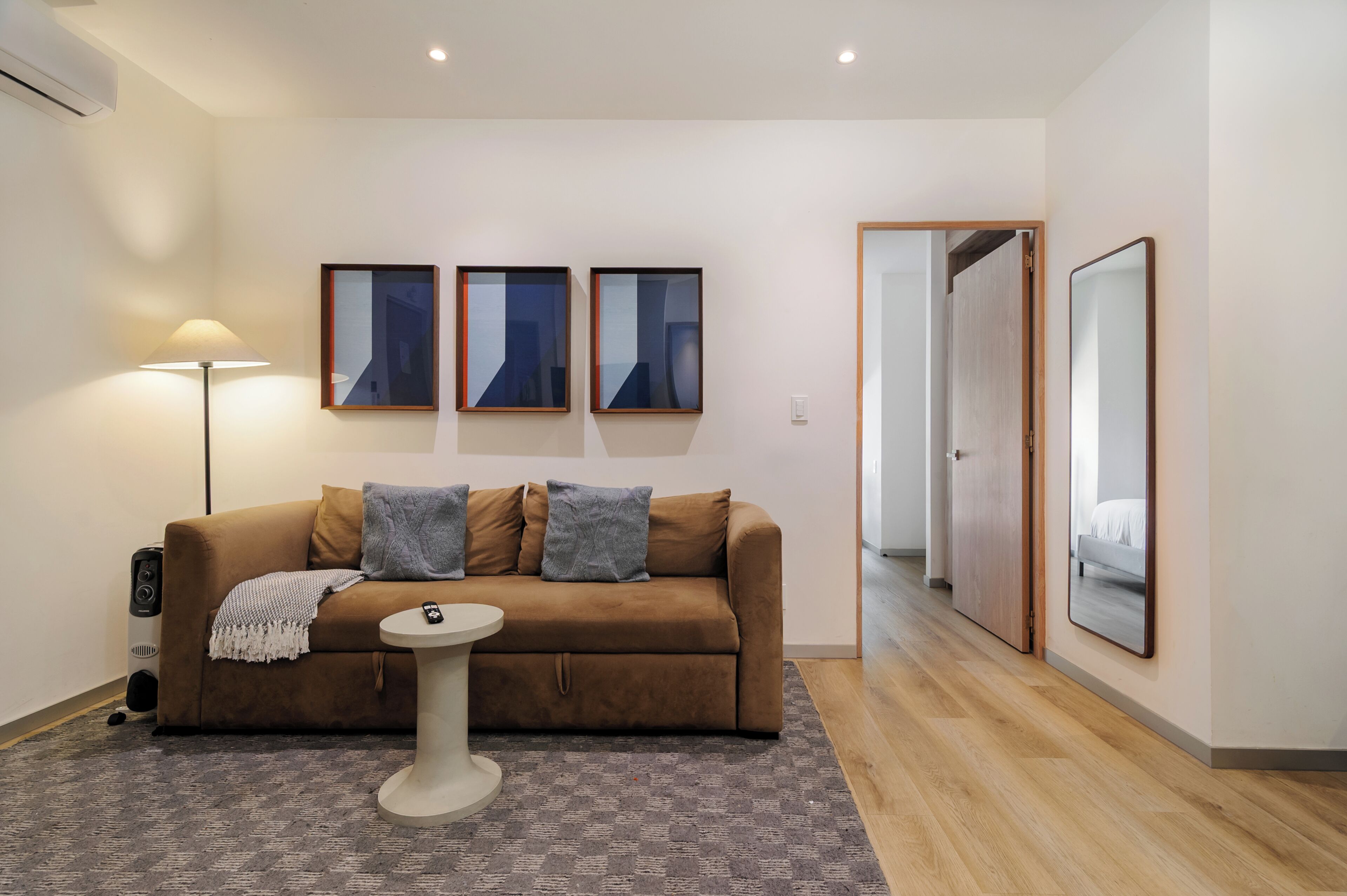 Un salon moderne avec un canapé marron, une petite table ronde et un lampadaire. Trois œuvres d’art encadrées ornent le mur, ainsi qu’une porte donnant sur une autre pièce et un grand miroir.