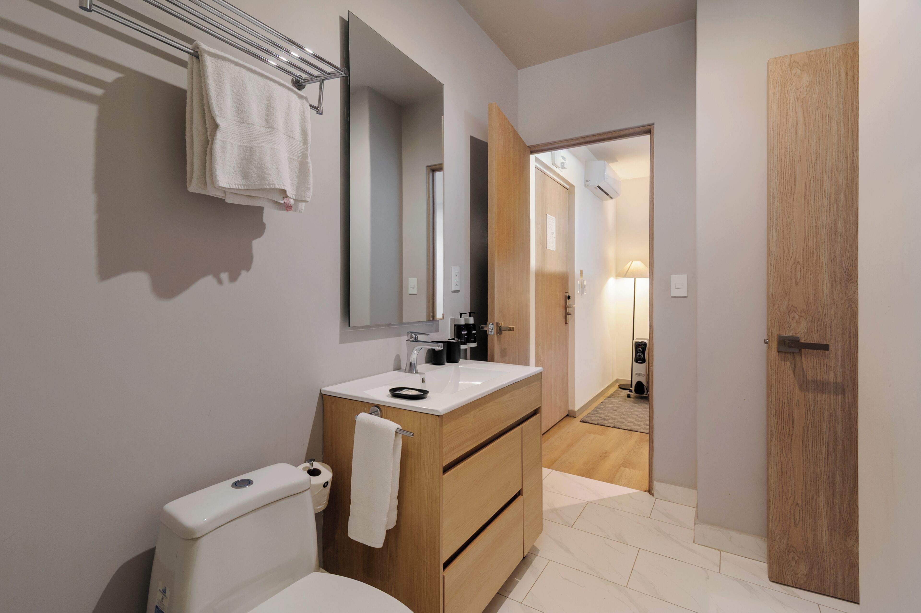 A modern bathroom with a toilet, sink, and mirror. There is a towel rack above the sink and a wooden door to the right.