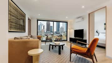 A modern living room with a sofa, a coffee table, and a TV. There is a large window with a city view.