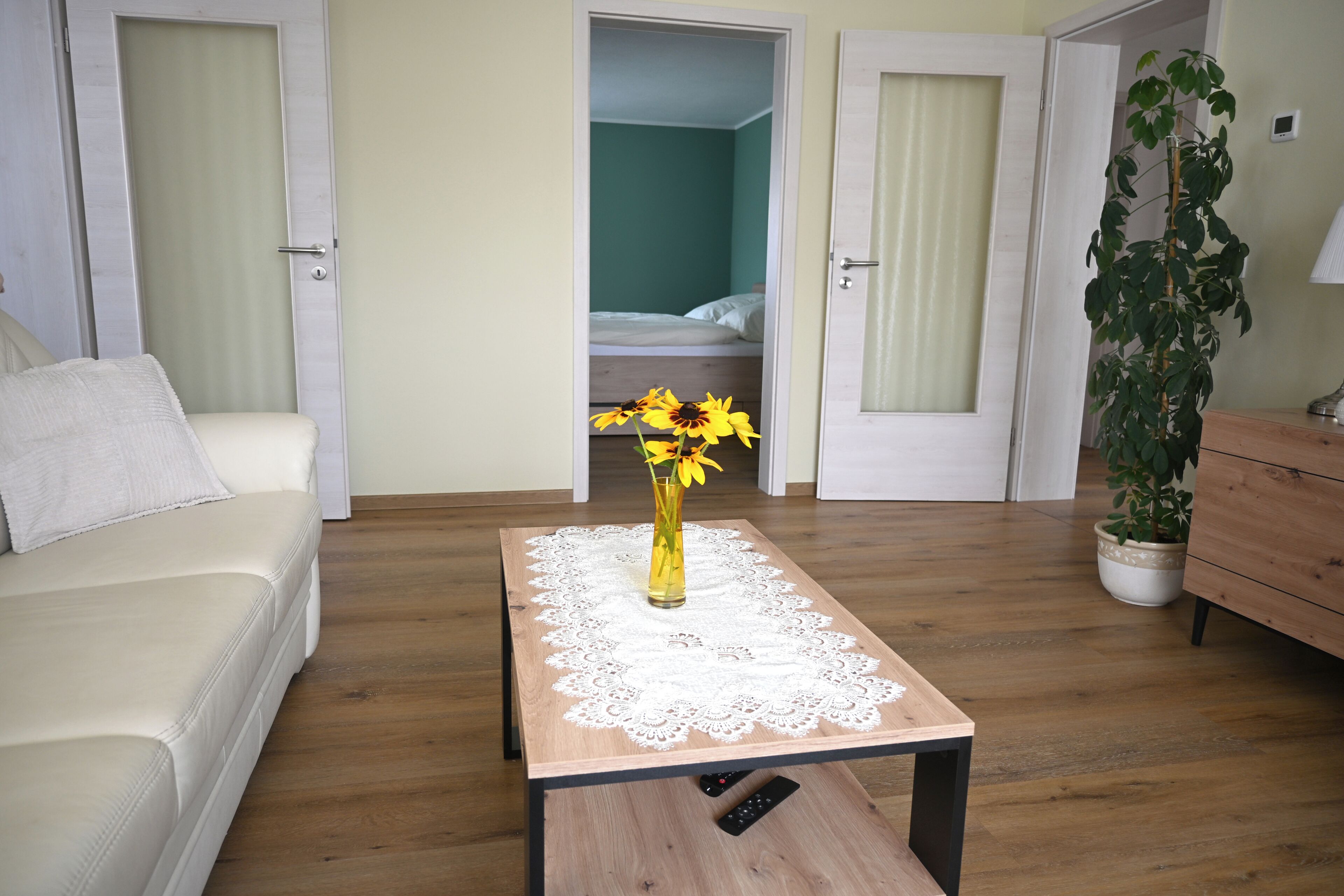 A hotel room with a wooden floor, a white sofa, a small table with a lace tablecloth, a vase with flowers, and a bed in the background.