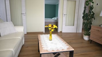 A hotel room with a wooden floor, a white sofa, a small table with a lace tablecloth, a vase with flowers, and a bed in the background.