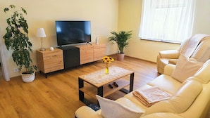 Uma sala de estar com sofá, mesa de centro, televisão, abajur e duas plantas em vasos.