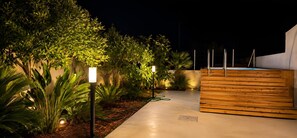 A nighttime outdoor area with a wooden bench, a concrete pathway, and various plants illuminated by lights. - Lemon Tree House con Piscina Privata Castelluzzo (San Vito Lo Capo)