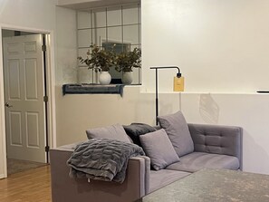 Ein Zimmer mit einem grauen Sofa, einem kleinen Tisch, einer Topfpflanze, einem Spiegel und einer Tür.