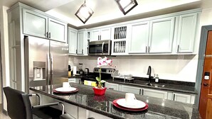 A modern kitchen with white cabinets, a stainless steel refrigerator, a microwave, and a sink. There are two black bar stools and a vase with flowers on the counter. - Enzo’s Suite Escape | Stylish 1BR in Liguanea (Kingston)