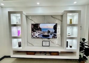 Ein Wohnzimmer mit einem großen Flachbildfernseher an einer weißen Wand, umgeben von Regalen mit verschiedenen Dekorationsobjekten.