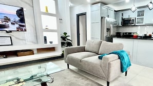 A modern living room with a glass coffee table, a beige sofa, and a kitchen area with cabinets and appliances. - Enzo’s Suite Escape | Stylish 1BR in Liguanea (Kingston)