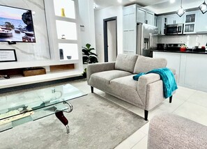 Ein modernes Wohnzimmer mit einem Glastisch, einem beigefarbenen Sofa und einer Küchenzeile mit Schränken und Geräten.