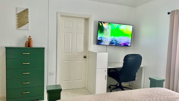 A bedroom with a bed, a desk, a chair, a dresser, and a television on the wall. - Enzo’s Suite Escape | Stylish 1BR in Liguanea (Kingston)