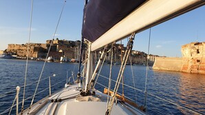 Het dek van een zeilboot met touwen en een mast, water, en een kustplaats met gebouwen.