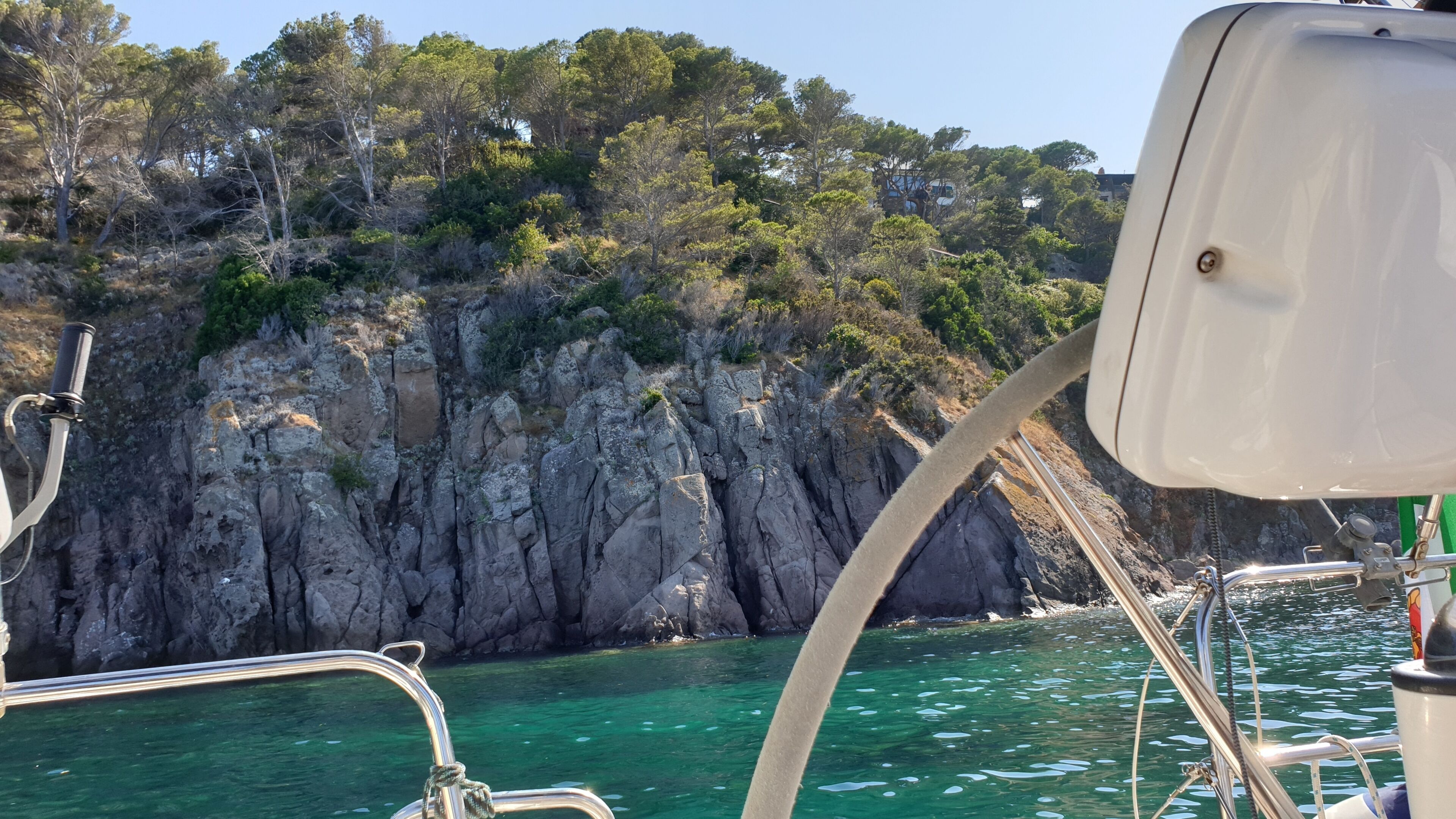 Una costa rocciosa con acque limpide, la ringhiera di una barca e una scogliera rocciosa con vegetazione.
