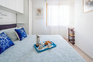 Ein ordentlich bezogenes Bett mit einem Frühstücksbrett, eine Wand mit einem gerahmten Kunstwerk und ein Fenster mit durchsichtigen Vorhängen.