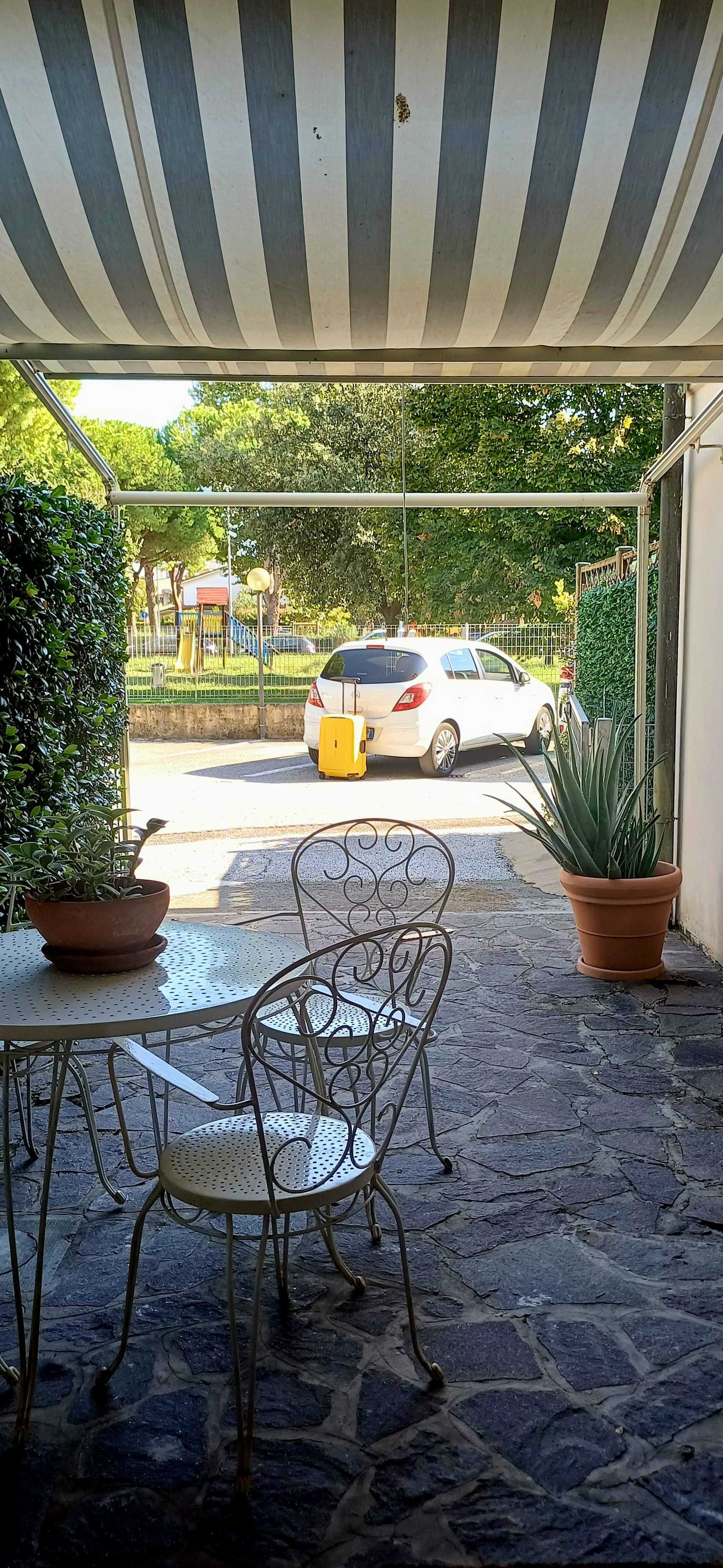 Un'area salotto all'aperto con sedie e tavoli in metallo, una pianta in vaso e vista su una strada con un'auto parcheggiata.