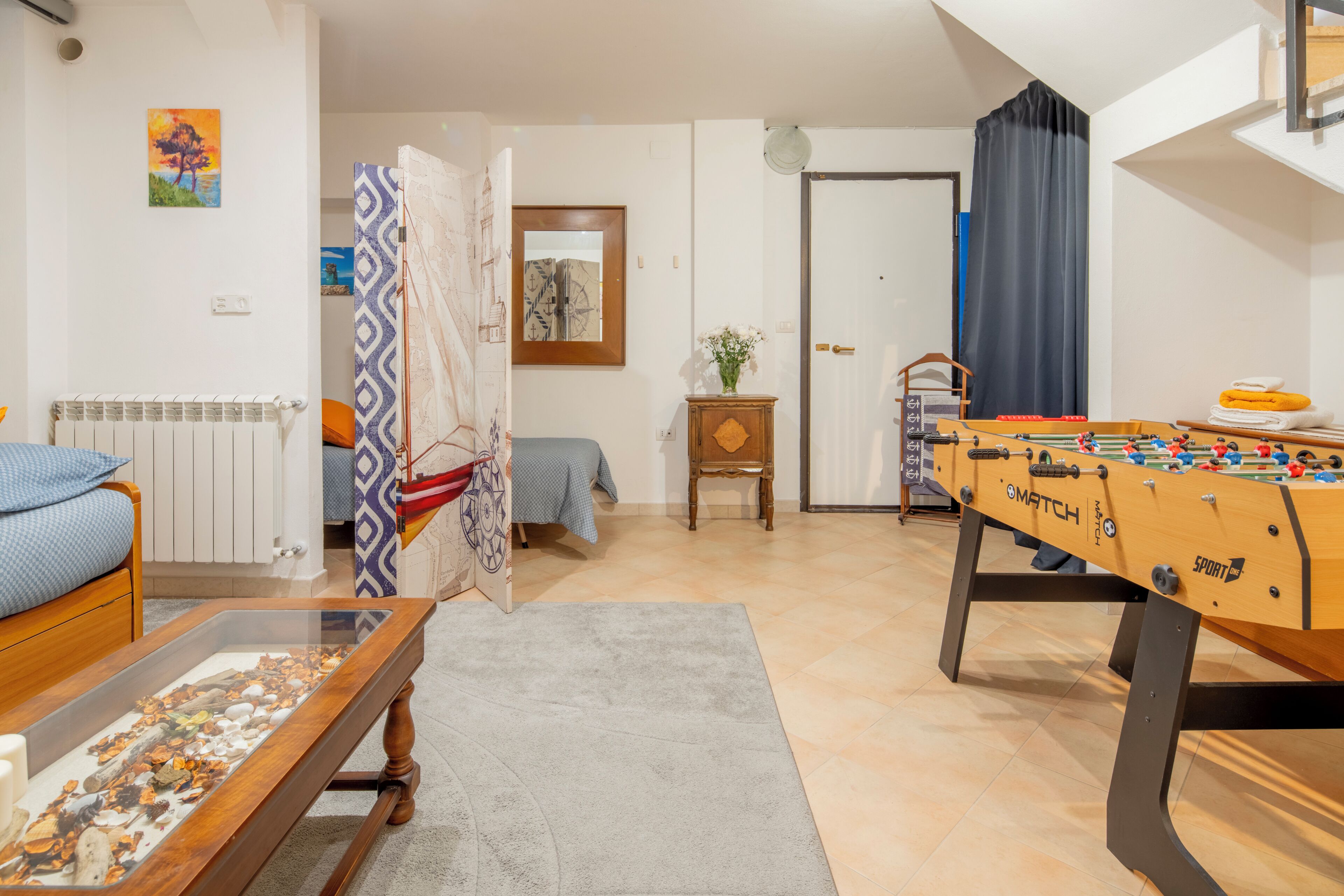 Ein gemütliches Wohnzimmer mit einem Kicker, einem Bett mit einem gemusterten Kopfteil, einem Heizkörper und einem dekorativen Glastisch mit Kieselsteinen.