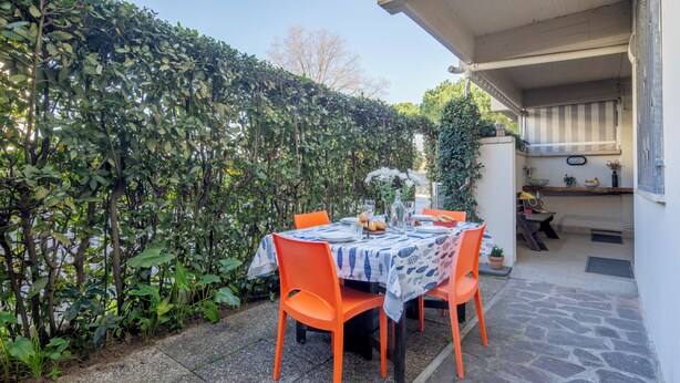 An outdoor dining area with a table set for two, surrounded by orange chairs, a stone floor, and a hedge. - A Tuscan Gem | Family Home with 2 Gardens & Private Parking (Viareggio)