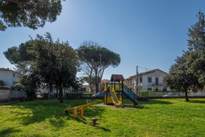 A playground with a slide and swings in a grassy area with trees and houses in the background. - A Tuscan Gem | Family Home with 2 Gardens & Private Parking (Viareggio)