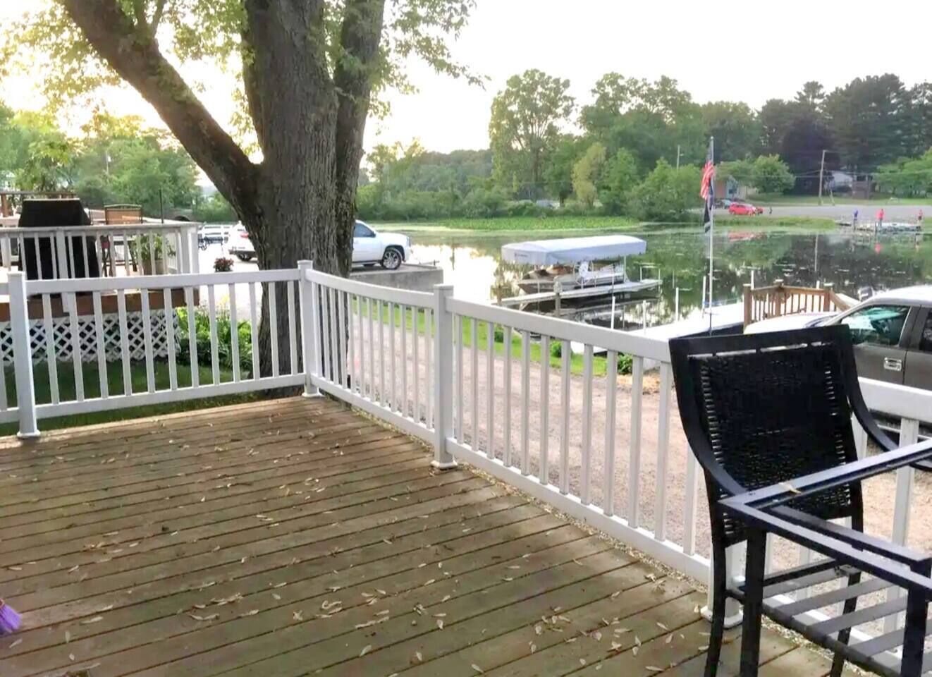 A wooden deck with a white railing, a black chair, and a view of a lake with boats and a dock.