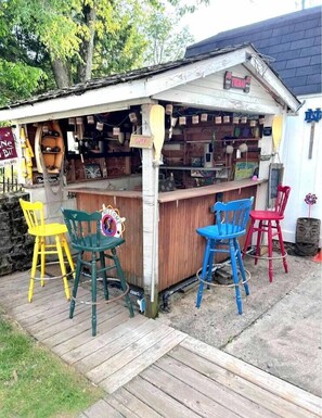 Un pequeño bar al aire libre con mostrador de madera, tres sillas coloridas y un letrero que dice 'OPEN'.