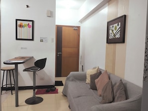 A small, furnished room with a sofa, a chair, a table, and a door leading to another room.