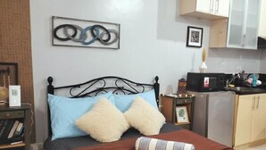 A bedroom with a bed, pillows, a bedside table, a nightstand, a desk, and a kitchen area with cabinets and appliances.