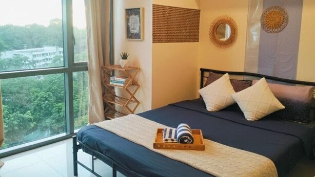 A bedroom with a large window, a bed with a blue mattress, two pillows, a bedside table with a lamp, and a view of greenery outside.