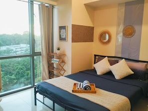 A bedroom with a large window, a bed with a blue mattress, two pillows, a bedside table with a lamp, and a view of greenery outside.
