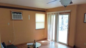 Sala de estar com ventilador de teto, uma mesa de vidro com um livro, sofá e uma porta de correr que dá para o exterior.