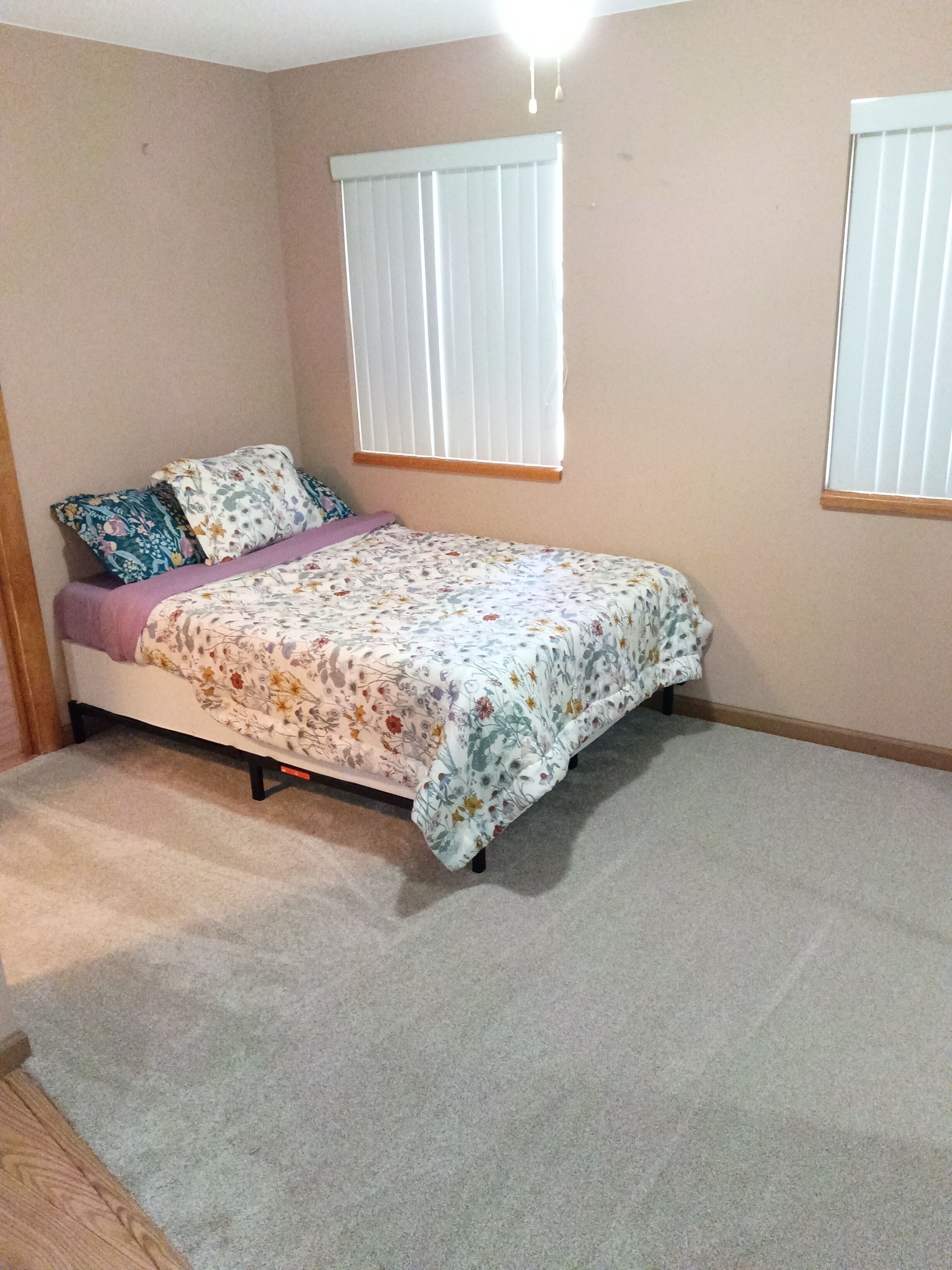 A bedroom with a bed, two windows with blinds, and a carpeted floor.