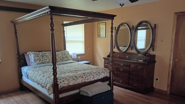 Uma cama com dossel acompanhada de um banquinho, uma cômoda com espelhos e uma janela com persianas.