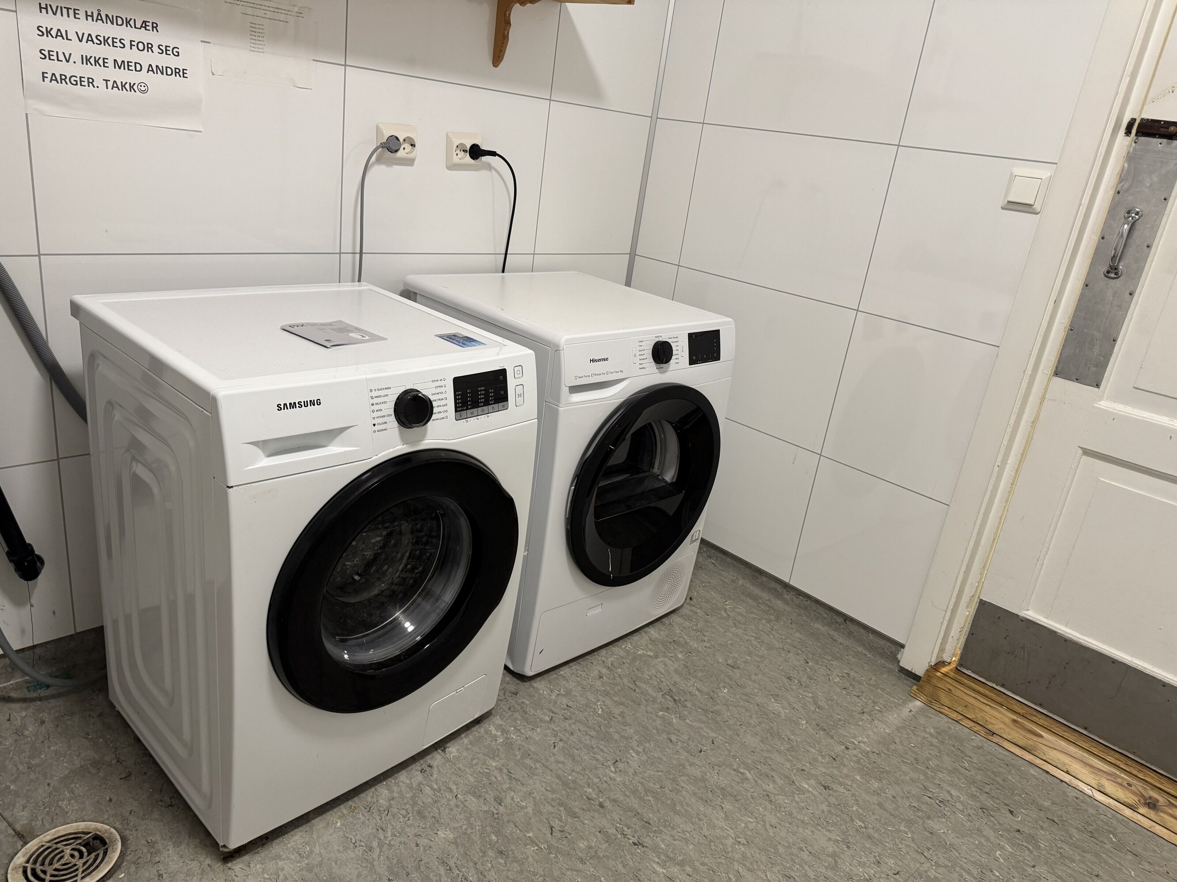 Dos lavadoras blancas de carga frontal en un lavadero con paredes de azulejos y una puerta de madera.