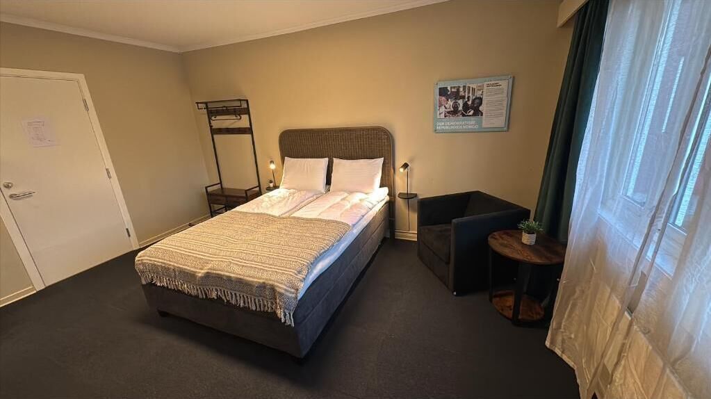 Habitación de hotel con cama, mesita de noche, silla, pequeña mesa, espejo y ventana con cortinas.