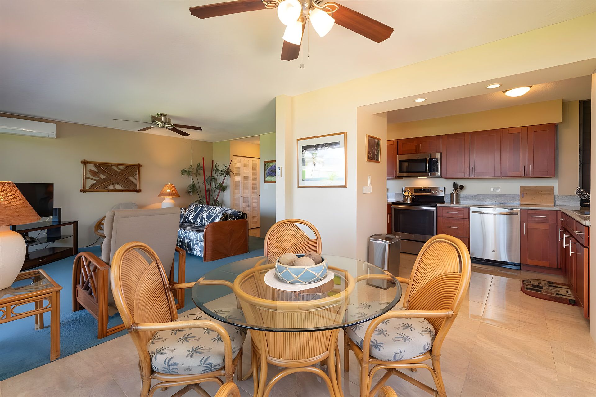 A modern living room with a glass dining table, wicker chairs, a ceiling fan, and a kitchen area with stainless steel appliances.