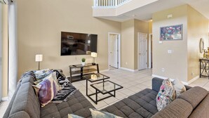A spacious living room with a high ceiling, a flat-screen TV mounted on the wall, a glass coffee table, and a sofa with multiple cushions. - 7 Bedroom Villa Near Disney World – Pool & Spa, Kissimmee (Kissimmee)