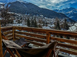 Eine Holzterrasse mit einem Stuhl und einer Tasche, mit Blick auf ein verschneites Dorf und Berge im Hintergrund.