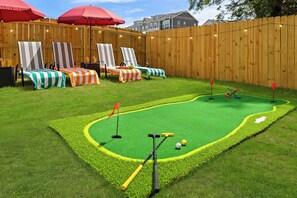 A backyard with a mini-golf setup, lounge chairs, and a wooden fence. - New! Sleeps 20 — Hot Tub, Minigolf, Firepit,Outdoor Movie & Game Room (Charlotte)