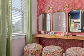 Une salle de bain avec un meuble-lavabo, deux tabourets à motifs, un miroir avec un cadre doré et un papier peint à fleurs.