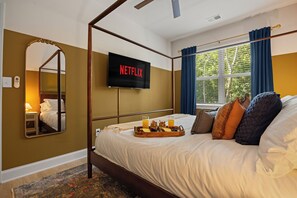 A hotel room with a four-poster bed, a flat-screen TV mounted on the wall, and a window with curtains.